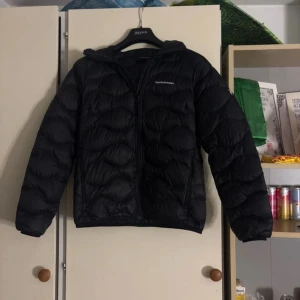 Svart dunjacka Peak Performance L - Svart dunjacka från Peak Performance i storlek L. Jackan har en snygg quiltad design, huva och dragkedja framtill. Perfekt för kyliga dagar och har en clean look som funkar till det mesta. Materialet är lätt och smidigt, med diskret logga på bröstet. Finns en defekt det hållet som är tejpat på bilden men dragkedjan funkar , passar även folk med storlek M
