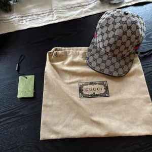 Gucci keps grå/blå  - Snygg grå/blå keps från Gucci med klassiskt GG-monogram i mörkblått över hela kepsen. Har en justerbar rem i svart skinn baktill och en röd-blå rand på sidan. Materialet är bomull och skinn, perfekt för dig som vill ha en lyxig och trendig accessoar.kepsen är i storlek L 59cm men eftersom man kan justera spänner så passar den typ alla 