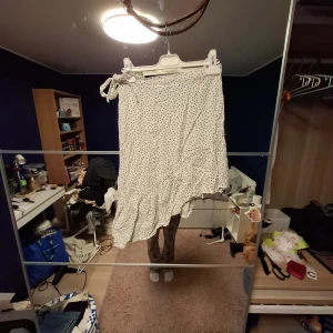 Vit prickig volangkjol från Kappahl - Supersöt vit kjol med svarta prickar från Kappahl. Kjolen har volangkant och asymmetrisk nederdel som ger en extra flowig look. Den har resår i midjan och knytband på sidan för en snygg detalj. Perfekt för dig som gillar lekfull stil.