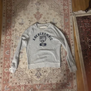 Grå sweatshirt Abercrombie & Fitch S - Grå sweatshirt från Abercrombie & Fitch i storlek S. Tröjan har ett tryck med tiger och texten 'Abercrombie 92' i marinblått framtill. Rund hals, ribbade muddar och långärmad. Materialet är mjuk bomull och polyester, perfekt för chill dagar.