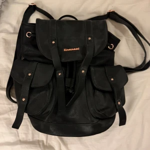 Svart ryggsäck  - Snygg svart ryggsäck i skinnimitation med två stora ytterfickor och snygga gulddetaljer. Invändigt har väskan ett svartvitt mönstrat foder och flera fack, perfekt för att hålla ordning på dina grejer.