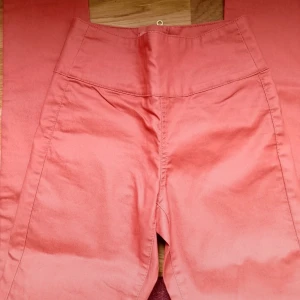 Högmidjade rosa byxor med dragkedja, strl. S/M - Strl. S/M. Ny med lapp. Snygga högmidjade byxor i en härlig rosa färg. Byxorna har raka ben, två bakfickor och en synlig dragkedja bak.  Perfekta för dig som vill sticka ut med färg och stil.