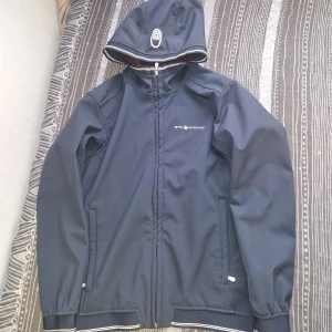 Mörkblå vindjacka från Sail Racing - Snygg mörkblå vindjacka från Sail Racing med huva och vit logga på bröstet. Jackan har dragkedja framtill, två sidofickor och ribbade muddar med vita ränder. Insidan är fodrad med mjuk fleece för extra komfort. Perfekt för blåsiga dagar.