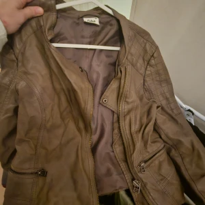 Brun skinnjacka från Cubus - Snygg brun skinnjacka från Cubus med quiltade detaljer på axlarna och dragkedjor vid fickorna. Jackan har en klassisk form och dragkedja framtill. Perfekt för dig som gillar en cool och stilren look.