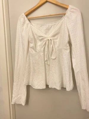 Vit blus med knyt och spetsdetaljer - Säljer en söt vit blus med långa ärmar och knyt framtill. Blusen har broderade spetsdetaljer och ett lätt utställt fall. Perfekt för dig som gillar romantisk stil och vill ha något luftigt och fräscht till garderoben.