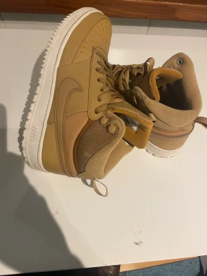 Nike Air Force 1 High Brun EUR 40 - Säljer ett par Nike Air Force 1 High i brun färg med vit sula. Skorna har snörning, högt skaft och är tillverkade i en mix av skinn och textil. Swoosh-loggan på sidan och klassisk rund tå. Perfekt för dig som gillar streetwear och vill sticka ut med snygga sneakers. använda en gång.