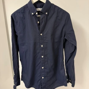 Mörkblå skjorta Tiger of Sweden - Snygg mörkblå skjorta från Tiger of Sweden i slim fit. Skjortan har klassisk button-down krage, vita knappar och lång ärm. Materialet är mjukt och känns som bomull, perfekt för dig som gillar stilrena och enkla plagg med bra passform. Storlek 39