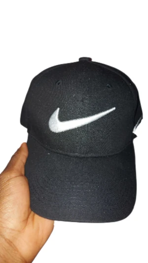 Svart Nike keps med vit nike logo  - Snygg svart keps från Nike med klassisk vit swoosh broderad framtill och på sidan. Justerbar rem med metallspänne där Nike-loggan är ingraverad. Insidan har Nike-tryck på svarta band. Tillverkad i bomull för skön passform.