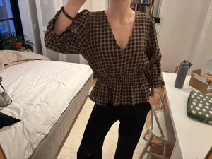 Brun och svart rutig blus med volang - Säljer en brun och svart rutig blus med volangkant nedtill och smockad midja. Blusen har lång puffärm och v-ringad fram, rundad hals bak. Materialet känns lätt och luftigt, perfekt för dig som gillar en chill men ändå snygg look. Snygg både åt ”rätt håll” och bakockfram! Kan inte hitta storlek men skulle säga s/m beroende på hur man vill att den ska sitta💕💕