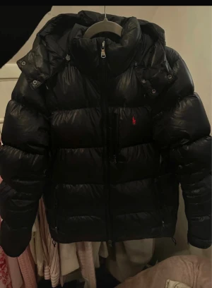 Marinblå pufferjacka från Polo Ralph Lauren - Säljer en svart pufferjacka från Polo Ralph Lauren med huva och dragkedja framtill. Jackan har en röd logga på bröstet, flera fickor med dragkedja och en riktigt fluffig look. 