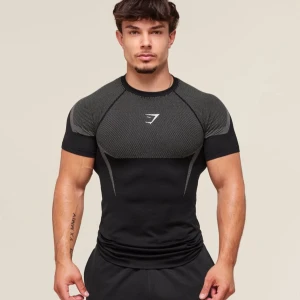 Gymshark Onyx 5 Black/Light Grey - Säljer en svart och grå tight träningstshirt från Gymshark med korta ärmar. T-shirten har sportig design med mönstrade partier på axlar och rygg samt Gymshark-logga på bröstet. Materialet är stretchigt och andas, perfekt för gymmet.