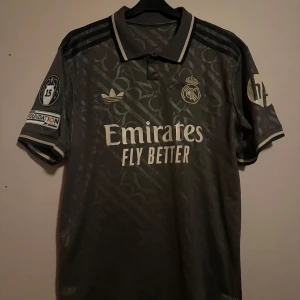 Grå Real Madrid Vini Jr. fotbollströja - Säljer en grå Real Madrid fotbollströja med Vini Jr. och nummer 7 på ryggen. Tröjan har korta ärmar, krage och snygga detaljer i silver. Adidas-logga och klubbmärke på bröstet, samt Champions League-märke på ärmen. 
