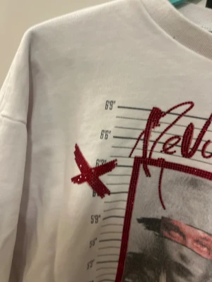 Vit Jeremy Meeks sweatshirt med röd glitterprint - Cool vit sweatshirt från Jeremy Meeks med stort tryck framtill, röd glittertext 'Revolution', och grafiska detaljer som höjdmätare och krona. Tröjan har rund hals och långa ärmar. Perfekt statement-piece för dig som gillar streetstyle.