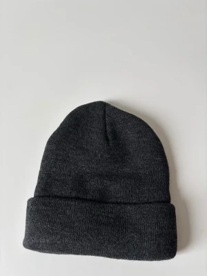 Mörkgrå stickad mössa - En enkel och stilren mörkgrå stickad mössa med uppvikt kant. Perfekt för kalla dagar och passar till de flesta outfits. Mjuk och skön känsla, klassisk form utan detaljer eller mönster.