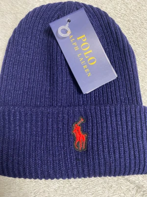 Marinblå ribbstickad mössa Polo Ralph Lauren - Snygg marinblå ribbstickad mössa från Polo Ralph Lauren med klassisk röd broderad logga framtill. Mössan är i mjukt material och har en uppvikt kant för extra stilpoäng. Perfekt accessoar för kyliga dagar och en ikonisk detalj till din outfit.