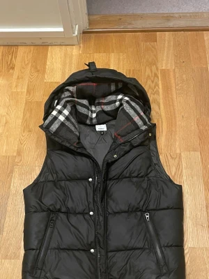 Svart dunväst från Burberry - Svart dunväst från Burberry med huva och klassiskt rutigt foder i grått, vitt och rött. Västen har dragkedja och tryckknappar framtill samt två fickor med dragkedja. Märkeslogga på vänster axel. Perfekt för kyliga dagar.