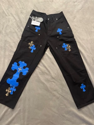 Chrome Hearts x Comme des Garçons jeans - Svarta byxor med blå och leopardmönstrade korsapplikationer i skinn och metalliska detaljer. Raka ben och flera fickor, med tydliga streetwear-vibbar. Märkestaggar från Chrome Hearts och Comme des Garçons. Perfekt för dig som vill sticka ut.