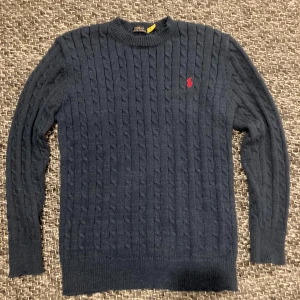 Mörkblå stickad tröja Polo Ralph Lauren - Snygg mörkblå stickad tröja från Polo Ralph Lauren med klassiskt kabelstickat mönster och rund hals. Tröjan har lång ärm och den ikoniska röda Polo-loggan broderad på bröstet. Perfekt för dig som gillar stilren och tidlös design.