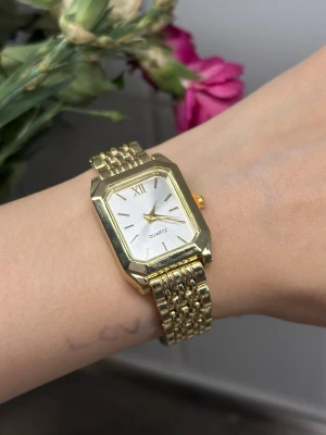 Guldfärgad fyrkantig quartzklocka - Snygg guldfärgad klocka med fyrkantigt ur och länkarmband. Urtavlan är silverfärgad med minimalistiska markeringar och romersk siffra vid tolv. Klockan har en klassisk och stilren design som passar perfekt till din outfit.