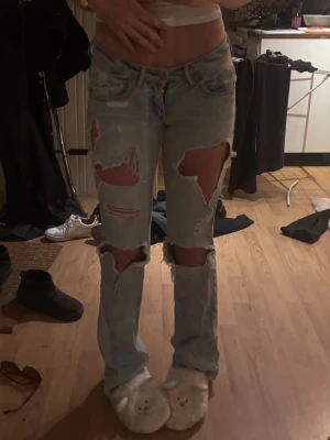Ljusblå lågmidjade jeans med hål - Säljer ett par ljusblå jeans med stora slitningar och hål på både fram- och baksida. Jeansen har låg midja och utsvängda ben, klassisk femficksmodell och är tillverkade i denim. Perfekta för en avslappnad och trendig look.