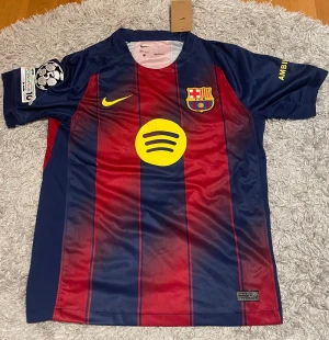 FC Barcelona Lamine Yamal tröja Nike S - Säljer en FC Barcelona matchtröja med Lamine Yamal och nummer 10 på ryggen. Tröjan är kortärmad, har klassiska blå och röda ränder, gula detaljer och sponsorloggor från Spotify och UNHCR. Tillverkad i lätt Dri-Fit polyester för maximal komfort. 