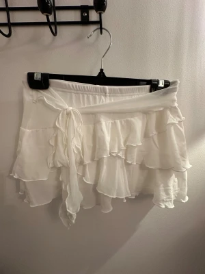 Vit volangkjol med knytband - Supersöt vit kortkjol med flera lager volanger och ett knytband i midjan. Kjolen har en flowy och lätt känsla, perfekt för dig som gillar romantisk och feminin stil. Materialet är tunt och mjukt, vilket gör den extra skön att bära. Passar dig som vill ha en fräsch och trendig look.