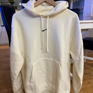 Vit hoodie från Nike med logga - Snygg vit hoodie från Nike med svart logga på bröstet och stor Nike-tryck på ryggen. Svarta band med Nike-tryck längs ärmarna, stor ficka framtill och huva med snörning. Perfekt för dig som gillar sportig och clean stil.