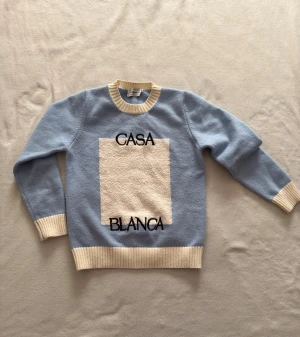 Blå stickad tröja Casablanca M - Snygg ljusblå stickad tröja från Casablanca med krämvita ribbade muddar och hals. Framtill finns en stor vit frottépatch med broderad text 'CASA BLANCA'. Perfekt för dig som gillar unika detaljer och cool design.