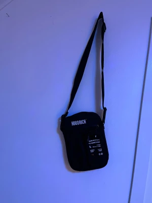 Svart axelväska från Hoodrich - Snygg svart axelväska från Hoodrich med justerbar rem och dragkedja framtill. Väskan har tryckt logga och text på framsidan, perfekt för att bära dina essentials. Stilren och enkel design som funkar till streetwear.