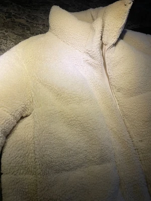 Vit teddyjacka med dragkedja - Mysig teddyjacka i vit färg med hög krage och hel dragkedja framtill. Jackan har en fluffig och mjuk yta i teddy-material som håller dig varm. Passformen är normal, perfekt för dig som gillar stilren och enkel look.