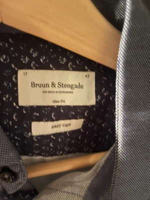 Slim fit grå skjorta Bruun & Stengade - Snygg grå skjorta från Bruun & Stengade i slim fit-modell. Skjortan har klassisk krage, svarta knappar och lång ärm med dubbla manschettknappar. Materialet är mjukt och lättskött bomull, perfekt för dig som gillar stilren och enkel design.