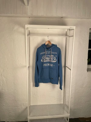 Blå hoodie från Superdry med tryck - Blå hoodie från Superdry med stort vitt tryck på bröstet och klassisk känguruficka. Tröjan har huva med snörning och ribbade muddar vid ärmslut och nederkant. Perfekt för chill dagar och streetstyle. Materialet är mjuk bomull och polyester. Finns några skavanker på höger arm och mage därav det billiga priset 