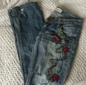 Blå jeans med broderade blommor - Snygga ljusblå jeans med broderade blommor och pärldetaljer längs ena benet. De har låg midja och bootcut, slitningar och hål på benen för en cool look. Har vissa slitningar vid hälarna. Midjemått 39cm och innerbenslängd 85cm. 