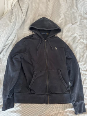 Svart hoodie från Polo Ralph Lauren - Svart hoodie från Polo Ralph Lauren med klassisk broderad logga på bröstet. Tröjan har huva med snörning, dragkedja framtill och två rymliga fickor. Tillverkad i mjuk bomull, perfekt för chill dagar. Långärmad och med normal passform.