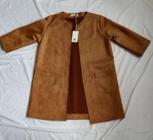 Beige mocka kappa från Pink Black - Snygg beige kappa i mockaimitation från Pink Black med rak passform och trekvartsärmar. Jackan har två stora fickor framtill och är öppen utan knappar eller dragkedja. Perfekt för dig som gillar stilrena och minimalistiska plagg.