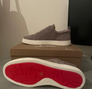Grå mocka sneakers från Louboutin - Snygga grå sneakers från Louboutin i mjuk mocka med vit platt sula och ikonisk röd undersida. Modellen har rund tå, snörning och diskreta metalldetaljer vid sidan. Perfekt för dig som vill ha en stilren och lyxig look med en twist.