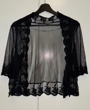 Svart spetsig bolerotopp från Vila - Säljer en svart bolerotopp från Vila i storlek M. Toppen är gjord i tunn mesh med vackra spetsdetaljer längs kanterna och nederdelen. Den har trekvartsärm och är öppen framtill, perfekt för att lägga över en klänning eller linne. Superfin och ger en cool vibe!