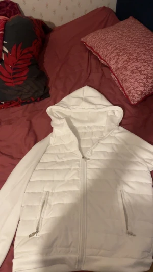 Vit quiltad pufferjacka med huva - Säljer en stilren vit pufferjacka i storlek M med quiltad design och huva. Jackan har dragkedja framtill och två fickor med dragkedja. Perfekt för dig som vill ha en clean och modern look under kyligare dagar.