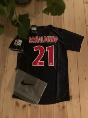 PSG Ronaldinho 21 Nike fotbollströja M - Säljer en klassisk Paris Saint-Germain fotbollströja från Nike med Ronaldinho och nummer 21 på ryggen. Tröjan är mörkblå med röd vertikal rand, PSG-märke på bröstet och Opel som sponsor. Korta ärmar och tillverkad i lätt, glansigt funktionsmaterial.