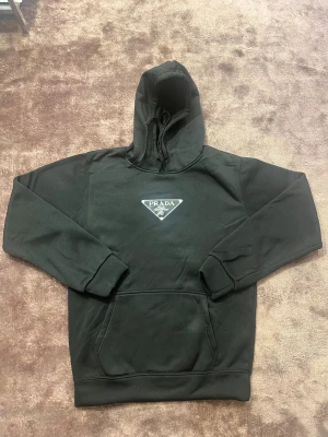 Svart hoodie från Prada med logga - Svart hoodie från Prada med ikonisk trekantig logga på bröstet. Tröjan har huva med snörning, långärmade ärmar och en stor magficka framtill. Materialet är mjukt och känns lyxigt, perfekt för dig som gillar designerplagg med clean look.
