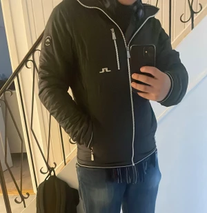 Svart quiltad pufferjacka J.Lindeberg - Svart pufferjacka från J.Lindeberg med huva, quiltad design och silvriga dragkedjor. Jackan har logotyp på bröstet och ärmen, samt praktiska fickor med dragkedja. Materialet är syntet och jackan har en normal passform. Perfekt för kyliga dagar.