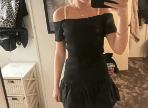 Svart offshoulder kortklänning - Supersnygg svart offshoulder klänning med smockad överdel och volang nedtill. Kort modell som sitter tight över kroppen och har en feminin siluett. Perfekt för dig som gillar trendiga och stilrena plagg med lite extra edge. Endast provad 