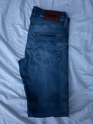 Blå jeans från Jack & Jones - Snygga blå jeans från Jack & Jones med klassisk femficksdesign och ljusa tvättade detaljer. Jeansen har normal passform och raka ben, med en brun läderpatch bak i midjan. Perfekta för dig som gillar stilrena och tidlösa jeans.