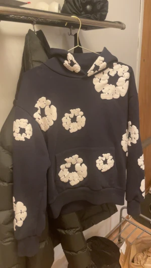 Mörkblå hoodie från Denim Tears S - Säljer en mörkblå hoodie från Denim Tears i storlek S. Tröjan har vita blommiga tryck över hela och en stor känguruficka framtill. Klassisk huva och mjukt material som känns riktigt skönt. Perfekt för dig som vill sticka ut med en unik streetwear-look.