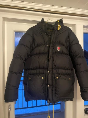 Svart dunjacka från Fjällräven XS - Svart dunjacka från Fjällräven i storlek XS. Jackan har huva, gul snörning och två stora fickor med knappar framtill. Klassisk Fjällräven-logga på bröstet och insidan. Den är i väldigt fint skick utan några defekter. Garanterat snabbt svar på frågor, funderingar och bud :)