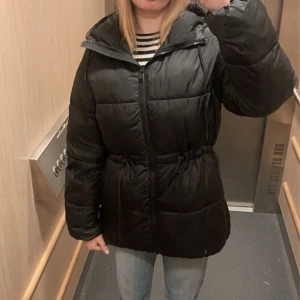 Svart pufferjacka med snörning - Säljer en svart pufferjacka med dragkedja och snörning i midjan. Har ett litet hål vid ena fickan!