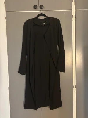 Svart lång kappa med knyt från 157 - Svart långkappa från 157 i storlek XS. Kappan har långa ärmar och ett snyggt knytband i midjan som ger en cool siluett. Enkel och stilren design, perfekt för dig som gillar minimalistiskt mode.