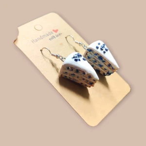 Blåbärs tårtbitar öronhänge  - Unika handgjorda örhängen formade som små tårtbitar med vit glasyr och blå detaljer. De har en cool och lekfull look, perfekta för dig som gillar att sticka ut. Örhängena är gjorda i polymerlera och har silvriga krokar. Perfekt accessoar för att ge din stil en rolig twist.