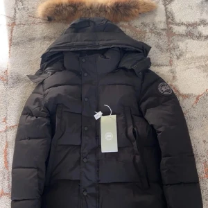 Svart dunjacka från Canada Goose - Svart dunjacka från Canada Goose med stor huva och snygg pälsdetalj. Jackan har flera fickor framtill, tryckknappar och dragkedja. Hängugglan är trasig annars perfekt Märkespatch på ärmen och insida i nylon. Perfekt för kalla vinterdagar och riktigt stilren look. Skriv om ni vill ha fler bilder tex på ugglan