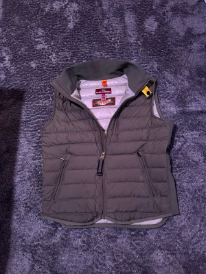 Mörk grön dunväst från Parajumpers YS - Snygg mörk grön dunväst från Parajumpers i storlek YS. Västen är quiltad med horisontella sömmar, har hög krage, tre dragkedjefickor och cool gul detalj vid halsen. Perfekt för lager-på-lager och har klassisk Parajumpers-logga bak i nacken.